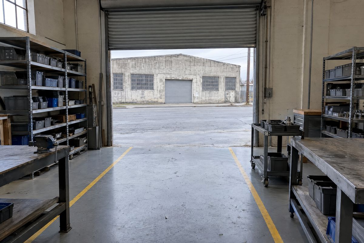 Workshop in an old concrete warehouse with a gray epoxy floor and yellow aisle lines, steel workbenches and shelving, and an open metal roll-up door.
