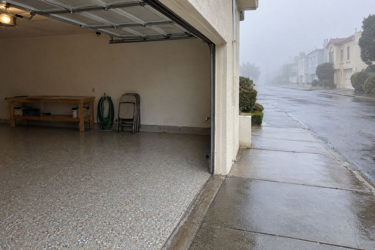 Street-level garage open to a foggy morning, its light gray, tan and white flake floor dry while the sidewalk outside is damp, with a workbench and hose.