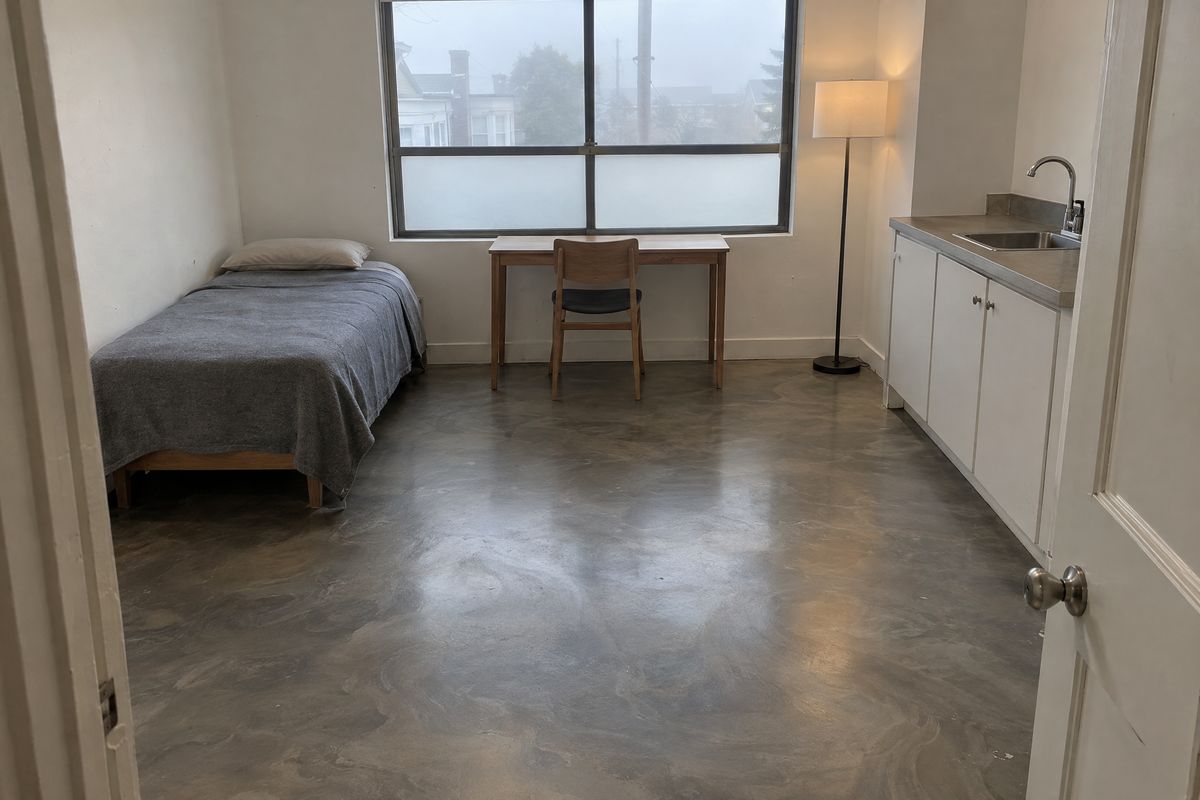 Garage converted into a small studio room with a muted pewter and warm gray metallic epoxy floor, a desk, a single bed, a kitchenette and a lamp.